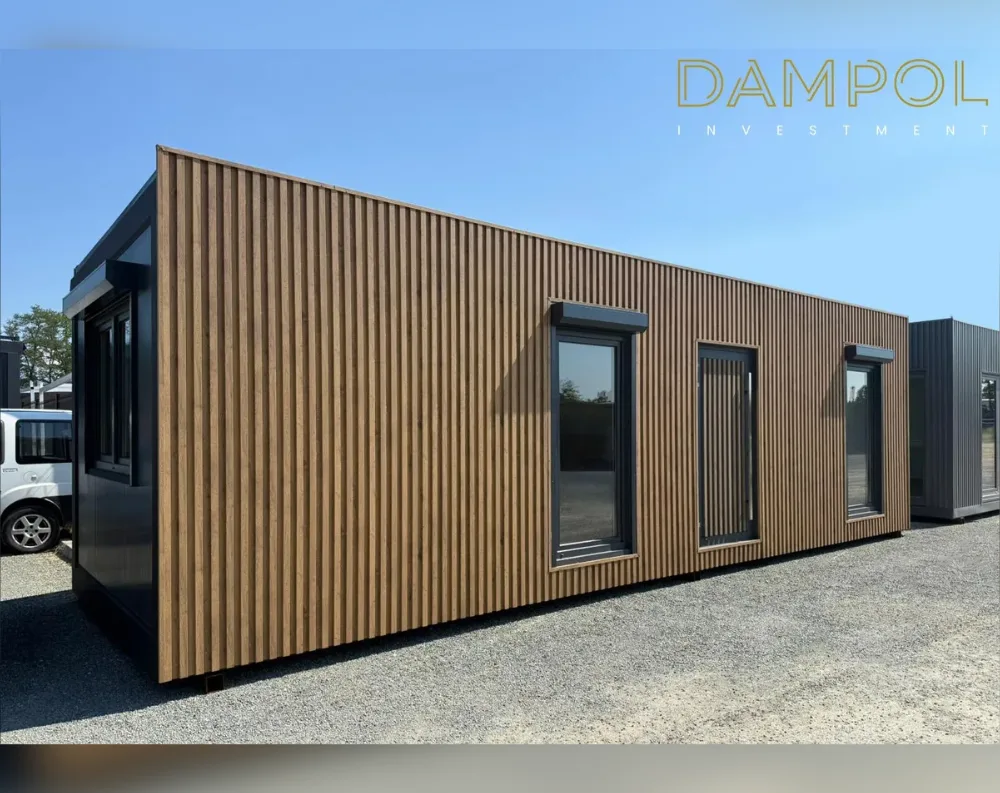 This modular building is a long, narrow rectangular structure with a flat roof, photographed at an angle that shows its full length receding into the distance. The exterior is clad in vertical wooden slats with a warm honey-brown finish, running uniformly across the walls and creating a textured, ribbed appearance. Dark charcoal-black trim frames the edges, roofline, and window surrounds, providing sharp contrast to the wood tones. Along the visible long side, there are two large windows and one narrower door positioned between them, all with black frames. The near end features another window on the black-paneled end wall. The building sits on light gravel, with a small black nameplate visible near its base.