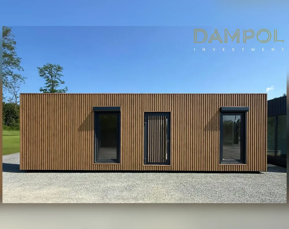 This modular building presents a long, low, rectangular volume with a flat roofline, giving it a sleek, horizontal profile. Its entire exterior is clad in vertical wooden slats in a warm honey-brown tone, creating a rhythmic, textured surface. The facade features three openings: on the left, a tall narrow window with dark charcoal framing; in the center, a matching-toned door with vertical slatted detailing that echoes the cladding, blending it almost seamlessly into the wall; and on the right, another tall dark-framed window mirroring the left. The structure sits on a gravel surface, set against a backdrop of green trees and clear blue sky, with another building's dark paneled edge visible at the far right.