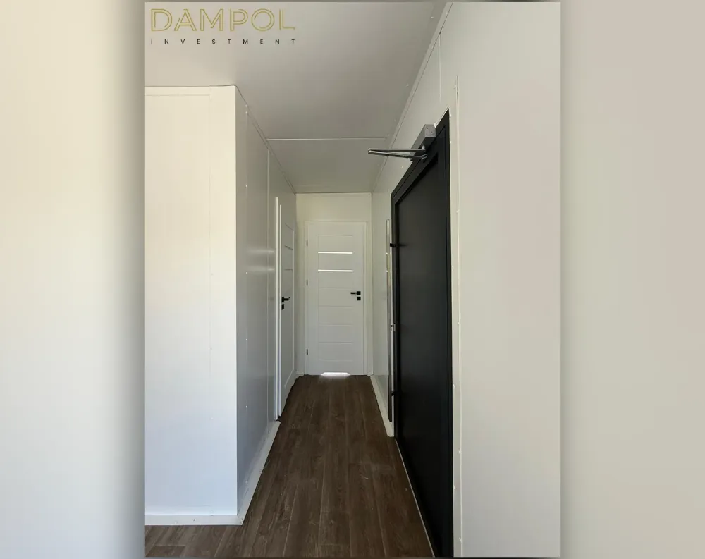 This photo shows the interior of a narrow hallway within the modular structure, rather than its exterior. The corridor features dark walnut-toned laminate flooring running its length, flanked by crisp white walls and ceiling. On the right side, a black door with a metal push-bar handle stands prominently in its white frame, suggesting a fire-rated or security entrance. Further down the hallway, a white paneled interior door with horizontal glass inserts and a black handle is visible, leading to another room. White built-in storage cabinets line the left wall. The ceiling is flat and painted white, with recessed lighting fixtures providing illumination throughout the narrow passage.