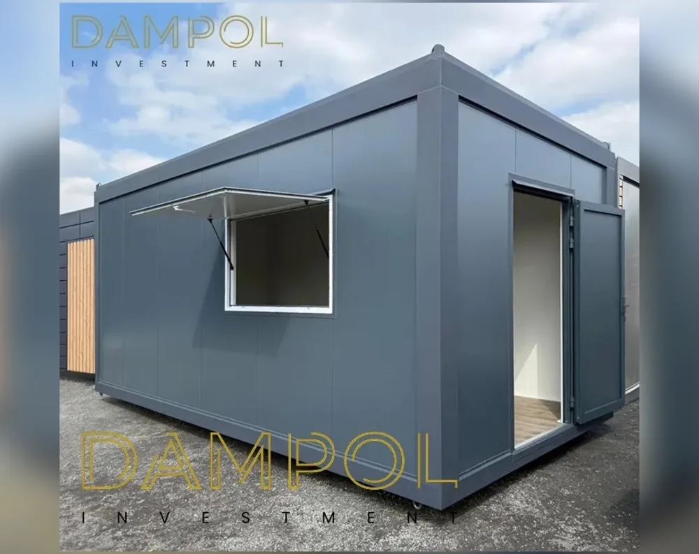 This modular building presents a sleek, rectangular form clad entirely in smooth, dark slate-grey composite panels with subtle vertical seam lines. The flat roof has a slightly raised edge, giving it a clean, boxy silhouette typical of contemporary garden offices or portable cabins. On the left side of the front-facing wall, a single square window features a hinged flap that opens outward and upward, propped by two support struts, revealing a plain white-painted interior beneath. To the right, a tall single door stands open, its grey exterior matching the cladding, exposing a bright white interior with light wooden flooring. The structure sits on a paved surface, with a wood-paneled building visible faintly in the background.