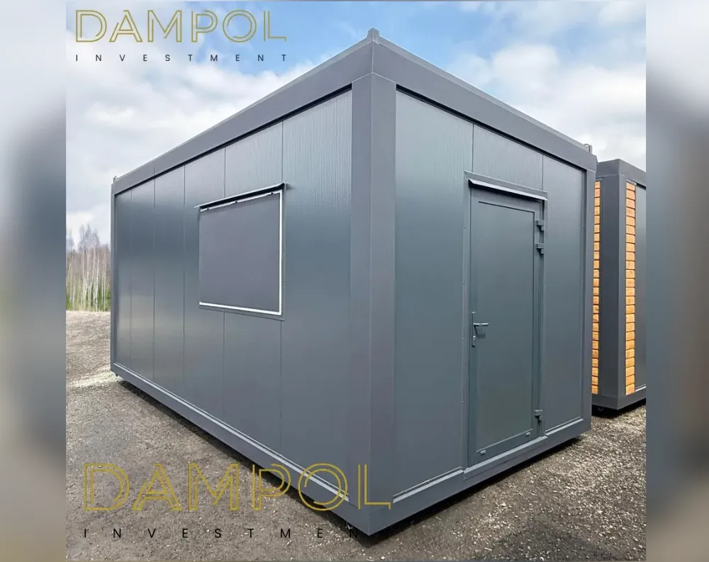 This modular building is a rectangular, single-story structure with a low-pitched, flat-style roof and dark charcoal-grey cladding featuring vertical ribbed paneling. Viewed from the corner, it appears compact yet solid, roughly proportioned like a small cabin or site office. On the shorter front-facing wall sits a single square window with a white or light-grey metal frame, positioned at mid-height. Adjacent to it, on the angled side wall, is a single steel door, painted the same dark grey, fitted with a metal handle and hinges, framed in a slightly lighter grey trim. The structure sits on a gravel surface, with another similar unit—clad in an orange-brown finish—visible just behind it.