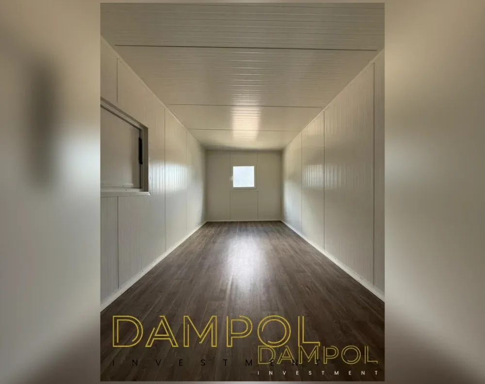 This photo shows the interior of a narrow, elongated modular room, viewed from one end toward the other, emphasizing its tunnel-like proportions. The walls and ceiling are finished in white ribbed paneling, creating a clean, uniform look with subtle horizontal grooves running along the surfaces. Natural light enters through a small square window centered on the far end wall, and another rectangular window is set into the left-hand wall partway down the room. The flooring is dark wood-toned laminate, contrasting sharply with the pale walls and adding warmth to the space. The ceiling is flat and unadorned, typical of a modular structural design, with no visible fixtures, giving the room a bare, ready-to-furnish appearance.