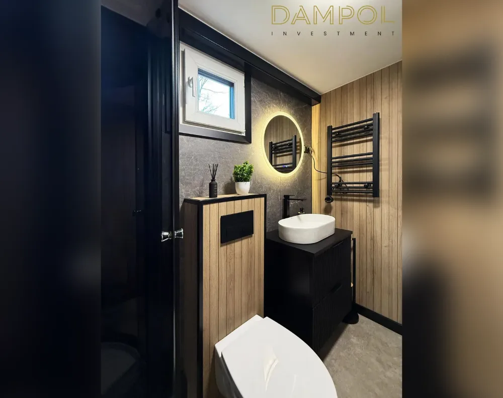 This compact bathroom features dark charcoal textured wall tiles paired with warm oak-toned wood paneling, creating a rich contrast throughout the space. A single white-framed window with black trim sits high on the left wall, allowing natural light in. Below it, a round backlit mirror glows warmly against the dark tile. A black vanity cabinet with vertical wood-paneled drawers holds a white rectangular vessel sink, accompanied by a matte black faucet. Two black ladder-style towel radiators mount on the wooden wall. A wall-hung toilet with a black flush panel sits beside a tall wood-and-black storage cabinet. The floor is polished dark grey concrete-style tile, and a black glossy door frames the entrance.