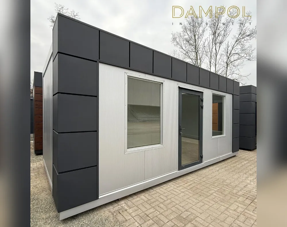 This modular garden office is a rectangular, single-story structure with a flat roof, photographed at an angle that shows both its short front face and long side elevation. The exterior combines two-tone cladding: dark charcoal grey vertical panels frame the corners, roofline, and edges, while the main wall sections are finished in a lighter grey ribbed panel material. The front facade features a large square window on the left, a full-height glazed door with dark metal framing in the center, and a smaller square window to the right. The building sits on a paved brick surface, with bare trees visible in the background against an overcast sky.
