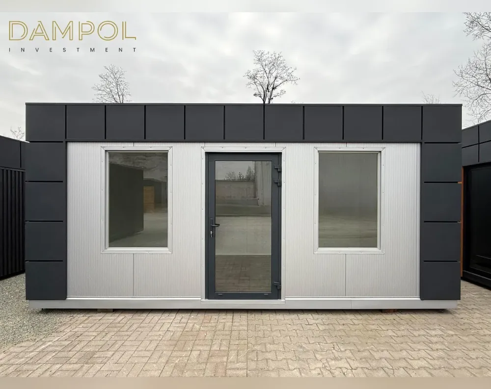 This modular unit is a long, low, rectangular structure with a flat roof trimmed in dark charcoal paneling. The main facade combines pale, ribbed light-grey cladding across the center with bold black vertical panel sections framing each corner, creating a strong contrast. A single glazed door sits centered, flanked symmetrically by two large rectangular windows with white frames, one on each side. The dark banding continues along the roofline as a fascia strip, echoing the black corner columns. The building rests on small support blocks above a paved brick surface, giving it a clean, elevated, boxy silhouette with crisp geometric lines and a minimalist, modern aesthetic overall.