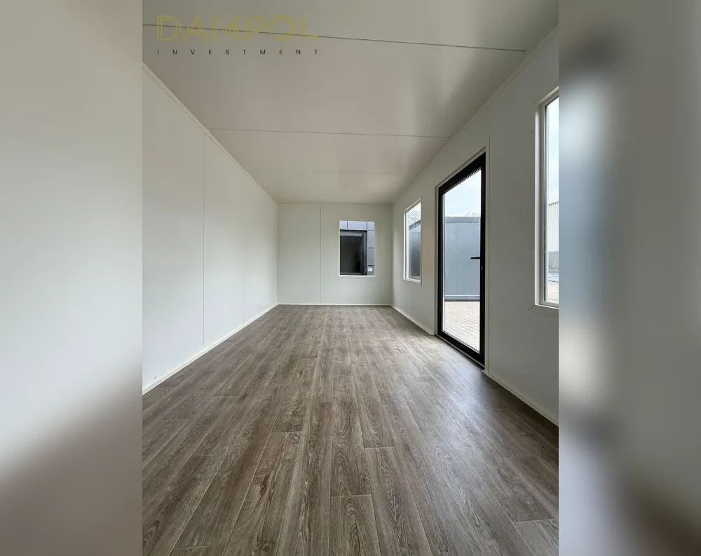 This is an interior view of a modular room with warm grey-brown wood-effect plank flooring stretching toward a glazed exit door at the far end. The walls and ceiling are finished in smooth white paneling, giving a clean, minimalist look. On the right side, two tall rectangular windows let in natural light, while a smaller square window sits recessed into the left wall near the rear corner. The full-height glass door, framed in dark grey or black, opens onto an outdoor paved area, with a glimpse of another structure visible outside. The room has a rectangular shape with a flat, low-pitched ceiling, and the overall aesthetic is streamlined, contemporary, and functional, well-suited to an office or garden studio setting.