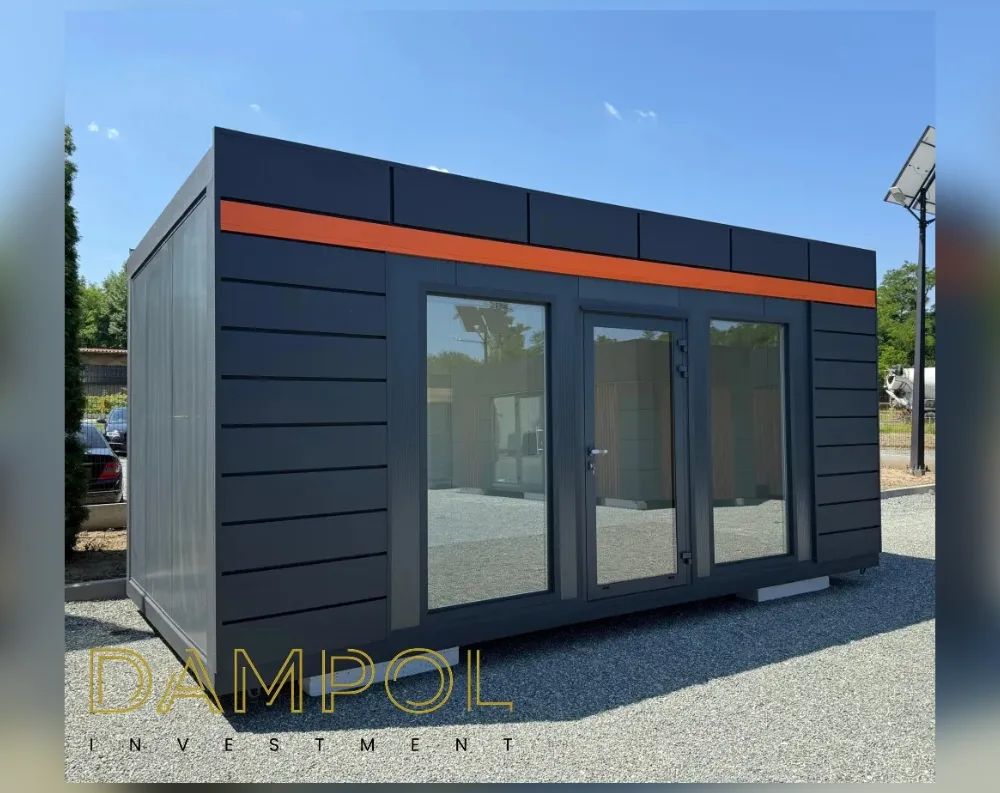 This modular garden office is a sleek rectangular structure with a flat roof, presenting a long, low profile with clean, boxy proportions. The exterior is clad in dark charcoal-grey horizontal slat paneling, giving it a modern, streamlined appearance. A bold orange horizontal stripe runs along the upper section of the front and side walls, just below the roofline, adding a striking accent to the otherwise monochrome façade. The front features a large glazed entrance: a central hinged glass door flanked by two fixed floor-to-ceiling window panels, all set within dark aluminum frames. The unit sits raised slightly off the gravel ground on visible support blocks, reinforcing its portable, prefabricated design.