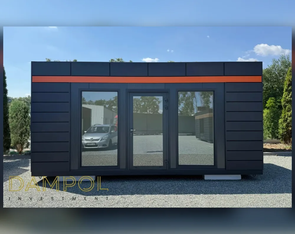 This modular building is a sleek, rectangular structure with a low-profile flat roof, wider than it is tall, giving it a horizontal, elongated silhouette. The exterior is clad in dark charcoal, almost black horizontal paneling, lending a modern, streamlined look. A bold orange horizontal stripe runs across the upper section, just below the roofline, adding a vibrant accent against the dark tones. The front façade features a symmetrical design: two large tinted glass windows flank a central glass-paneled door, all set within matching dark frames. The structure rests on small white support blocks, elevating it slightly off the gravel ground it sits on.