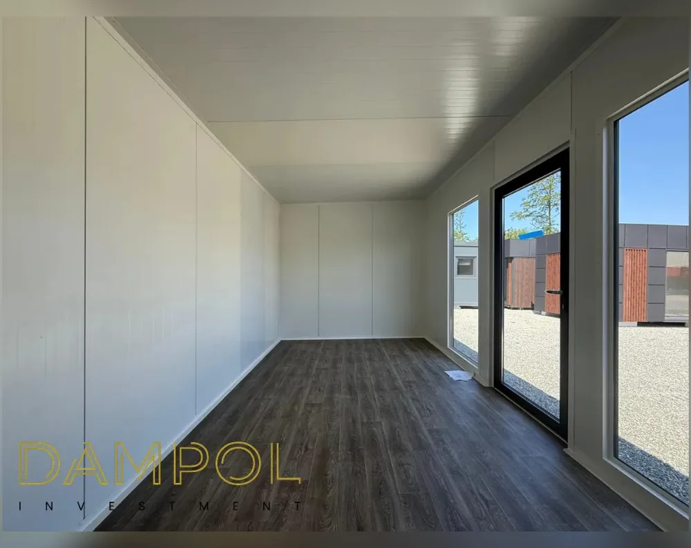 This photo shows the empty interior of a modular garden office, captured from a rear corner looking toward the entrance. The room is a simple rectangular box with a flat, low-profile ceiling finished in smooth white panels, matching the plain white wall cladding on the left and rear walls. The floor is laid with dark walnut-toned laminate planks running the length of the space. Along the right-hand wall runs a continuous glazed façade: two large fixed windows flank a central black-framed glass entrance door, all set within slim dark frames. Through the glazing, gravel ground and neighboring modular units with grey and wood-effect panelling are visible outside under a clear blue sky.