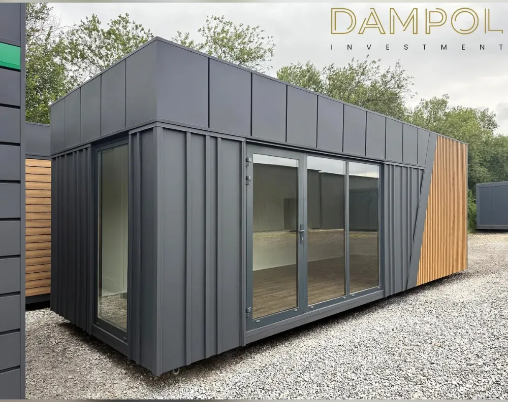 This modular garden office is a sleek, elongated rectangular structure with a low-profile flat roof, clad entirely in dark charcoal-grey vertical panelling that gives it a crisp, modern industrial look. On the short front-facing end, a tall floor-to-ceiling window sits beside a narrow vertical strip window near the corner. The long side features a wide glazed section comprising a hinged glass door paired with two sliding glass panels, allowing generous natural light into the interior. Toward the far end, the grey cladding gives way to a diagonal-cut accent panel finished in natural light wood slats, creating a striking contrast. The building rests on a gravel surface, with greenery visible behind it.