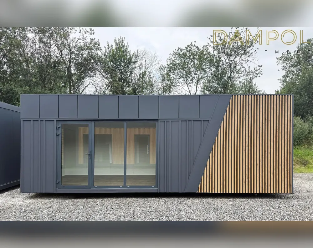 This is a sleek, rectangular modular building with a low, flat roofline, sitting on gravel ground before a backdrop of dense green trees. The structure is clad primarily in dark charcoal-grey paneling with vertical ribbed detailing, while the right-hand section features a striking contrast of natural light-wood vertical slats, divided diagonally from the grey by a bold angular line. The front facade is dominated by a wide glass entrance: a single hinged door with a full-length window pairs with a large sliding glass panel beside it, together spanning most of the wall and revealing a bright interior with wood-paneled walls. No additional windows appear elsewhere on this visible side, keeping the design minimal and modern.