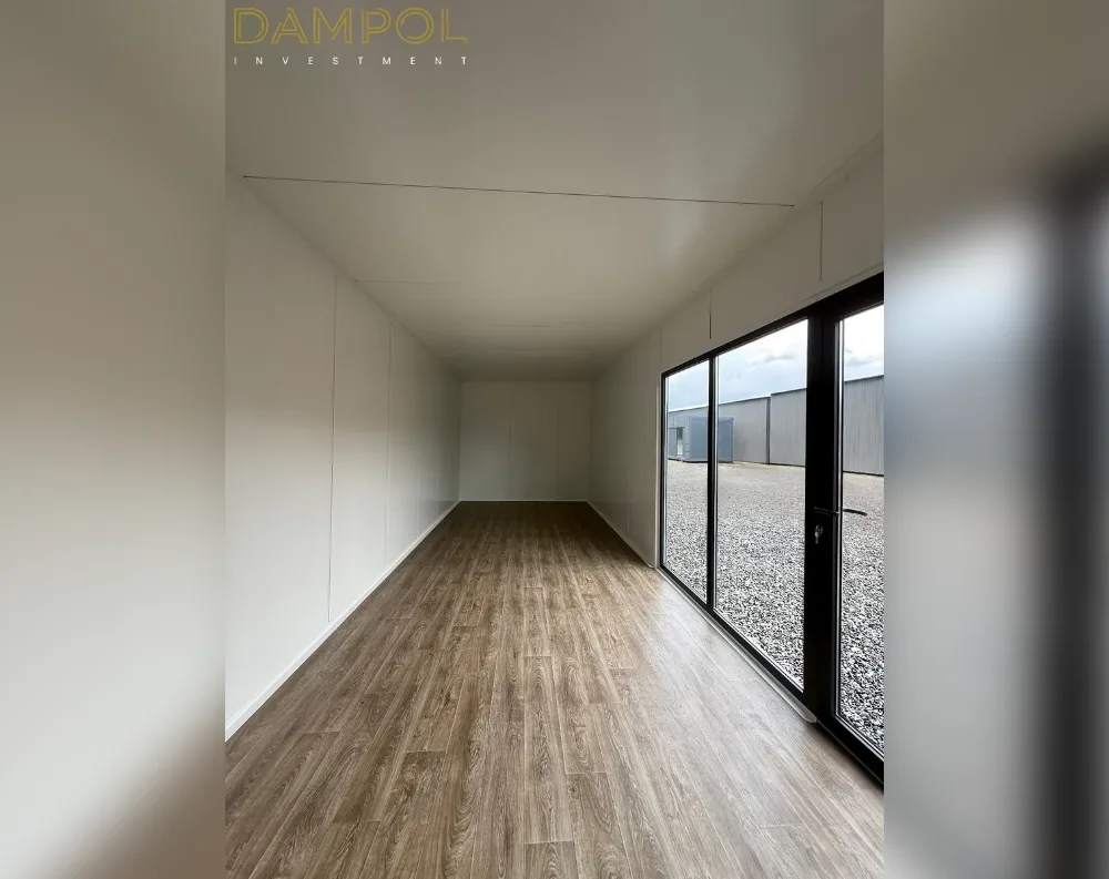 This is an interior view of a long, narrow modular room with a simple rectangular footprint stretching back several meters. The walls and ceiling are finished in smooth white paneling, uninterrupted except for a subtle seam line running across the ceiling. Warm, medium-brown wood-look laminate flooring runs the entire length of the space, adding contrast to the pale walls. At the far right end sits a black-framed glass door system with multiple large panes, including a hinged door with a visible handle, allowing daylight to flood in and revealing a gravel yard and a grey industrial building outside. No other windows are visible along the side walls.