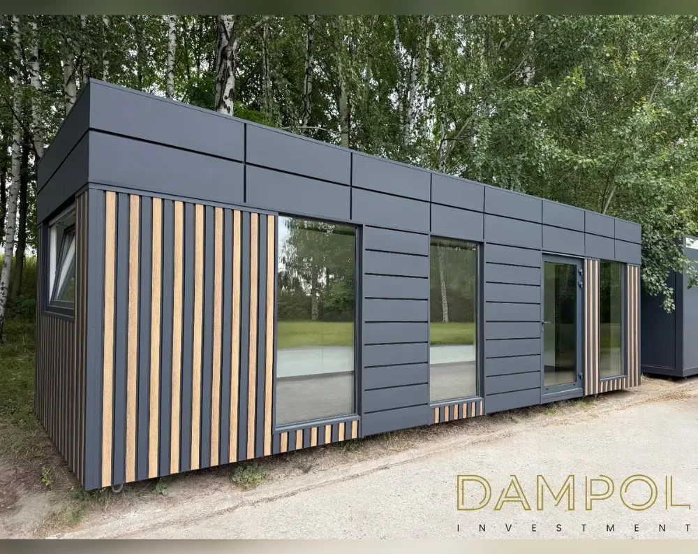 This modular garden building is a long, low rectangular structure with a flat roof, photographed at an angle that shows both its short end and long side. The exterior is clad in dark charcoal-grey panels, contrasted by vertical light wood-toned slats accenting the corners and framing sections of glazing. Along the front, there are three large fixed windows and one central glass entry door with a slim adjoining sidelight window, all set in dark aluminum-style frames. A smaller window sits on the shorter left-facing wall. The roofline is crisp and minimal, overhanging slightly. The building rests on the ground with visible support blocks beneath, set against a backdrop of birch trees and gravel.