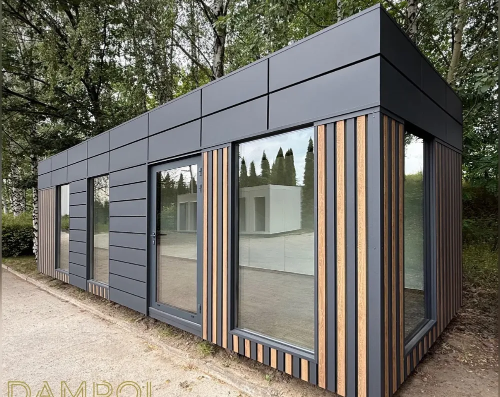 This modular garden office is a sleek, elongated rectangular structure with a flat roof, clad in dark charcoal-grey paneling arranged in horizontal ribbed strips. The building's corner is angled toward the viewer, showcasing two full glass walls that meet at this point. Vertical wood-toned slats frame the glazing, adding warmth against the dark exterior. Along the front, there are three large windows and one glass door with a slender frame, all positioned in a row. The corner section features expansive floor-to-ceiling glass panels, offering wide visibility inside. The structure sits on a gravel path, surrounded by tall trees, with another similar white modular unit visible in the reflection and background.