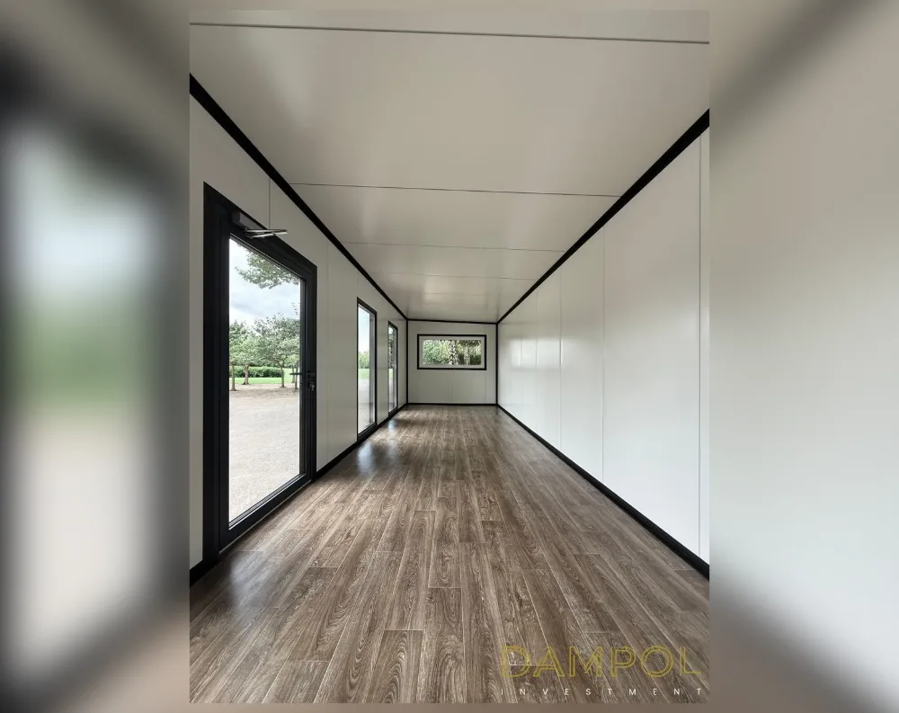 This photo shows the interior of a long, narrow modular building, likely a garden office or container-style structure, stretching into the distance with striking symmetry. The walls and ceiling are finished in smooth white paneling, framed by black trim along the seams and corners, creating a crisp contrast. The floor is laid with warm, dark wood-effect laminate planks running the length of the room. On the left side, a full-height glass door with black framing sits near the front, followed by a series of narrower vertical windows and one small horizontal window further down, all trimmed in matching black. The space is bright, minimalist, and evenly proportioned, with a flat ceiling and clean architectural lines throughout.