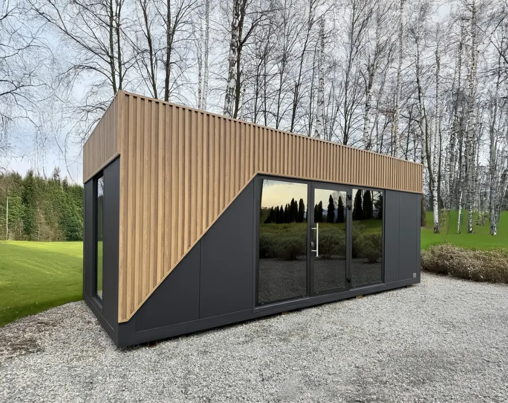 This modular garden building has a sleek rectangular form with a gently sloped mono-pitch roof, angled higher at the front and lower toward the back. The upper portion is clad in vertical light-oak wooden slats, while the lower half is finished in smooth charcoal-black panels, divided diagonally by a striking angled line where wood meets black. The front façade features full-height black-framed glass: a large fixed window on the left, a central double glass door, and another fixed window to the right. A smaller black-framed window sits on the visible side wall. The structure rests on a gravel pad, surrounded by manicured lawn and bare birch trees.