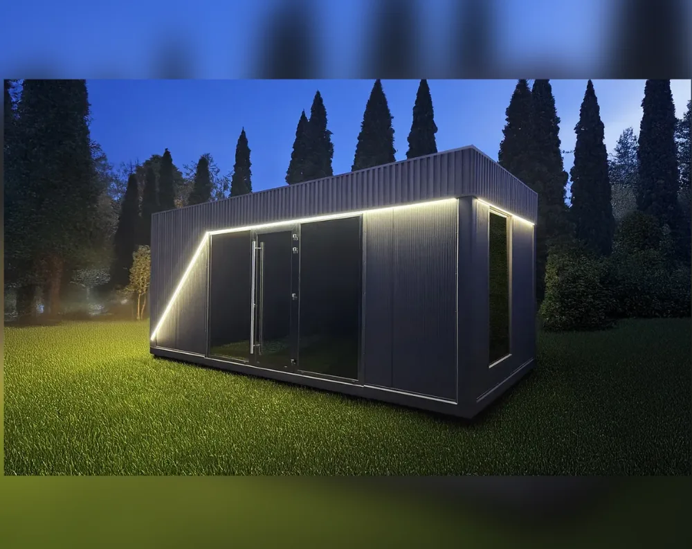 This modern garden office is a sleek rectangular structure with a low-pitched, slanting roof that angles upward toward the rear, clad in dark charcoal-grey corrugated paneling. The front facade features a full-height glass entry system: a diagonal angled window on the left, two large glass panels flanking a central glass door with a vertical handle, all trimmed in black frames. A strip of bright LED lighting runs along the underside of the roofline and down the angled corner edge, casting a warm glow. On the right side, a tall narrow window is set into the dark paneling. The building sits on a raised platform amid a manicured lawn, backed by tall silhouetted evergreen trees under a dusky blue sky.