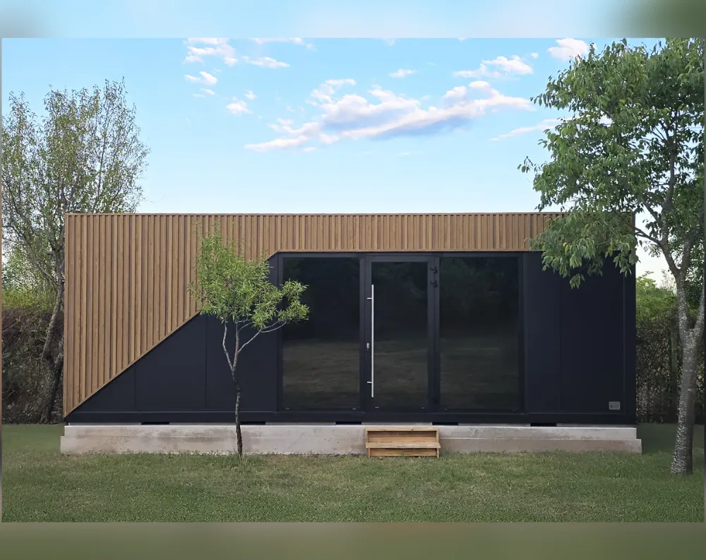 This modern modular building has a sleek, rectangular form with a flat roof line and a low-slung, horizontal profile. The upper portion is clad in warm, vertical wood slats that sweep diagonally down the left side, creating a striking angled accent against the matte black lower façade and side panels. Centered on the front is a large glass entrance system: a single black-framed door with a slim silver handle, flanked by two wide fixed glass panels that mirror its dark tint. Small wooden steps lead up to the door from the lawn. The structure sits on a raised concrete foundation, surrounded by grass and framed by trees on either side.