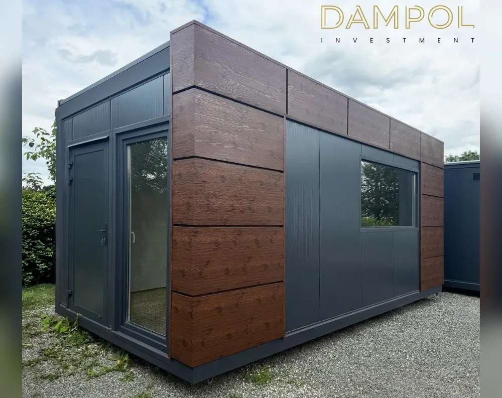 This is a compact, rectangular modular cabin with a flat roof, photographed at an angle that shows both its short front face and long side elevation. The exterior combines two materials: dark charcoal-grey paneling and warm brown wood-look cladding arranged in horizontal bands, meeting at crisp mitred corners. The front facade has a full-height grey door on the left paired with a tall glass panel beside it, while the long side features a smaller single door, another slim window, and one larger rectangular window further along. The structure sits on a gravel surface, with greenery and hedges visible to the left and another similar building partially visible on the right.