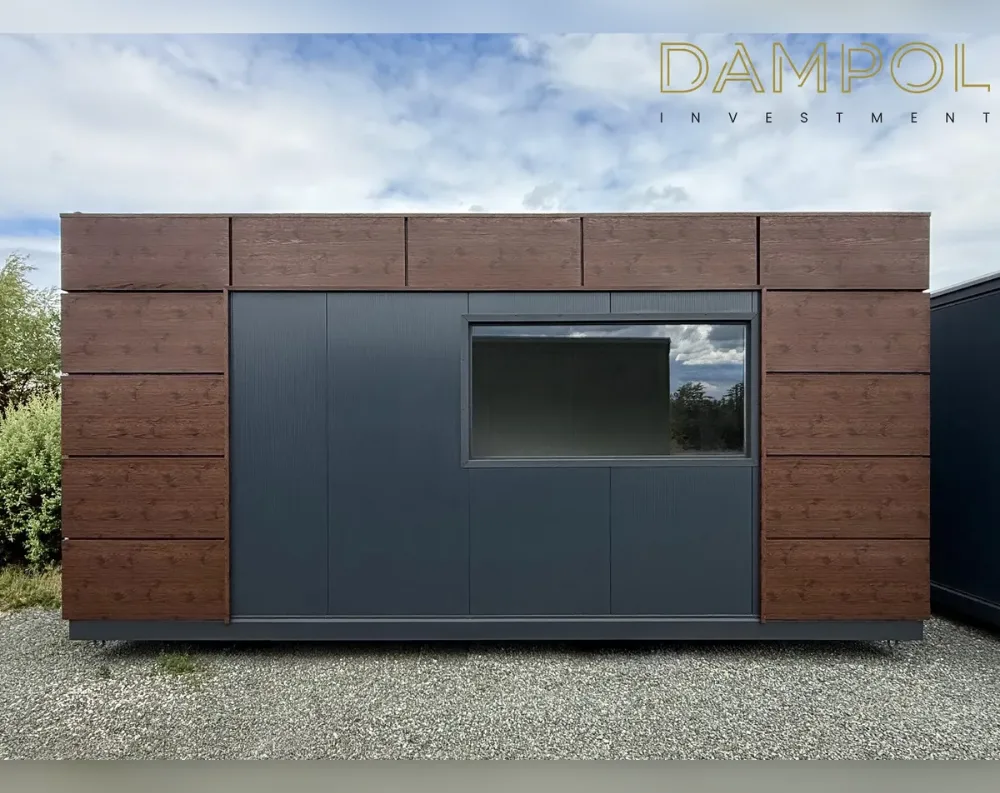 This modular garden building has a low, wide rectangular shape with a flat roof, giving it a sleek, contemporary profile. The structure combines two cladding materials: rich, dark walnut-toned wood paneling wraps the upper band and flanks both sides, while the central façade is finished in smooth charcoal-gray composite panels. Centered in this dark section is a single large horizontal window with a slim black frame, positioned slightly right of center, reflecting the surrounding trees and sky. No visible entry door appears on this front elevation, suggesting access may be from another side. The building sits on a raised base with small support feet, elevating it slightly above the gravel ground below.