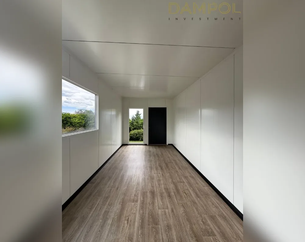 This is an interior view down the long, narrow corridor of an elongated modular building, its proportions emphasizing depth over width. The walls and ceiling are finished in smooth, matte white paneling with visible seam lines marking the modular sections. Warm grey-brown wood-effect vinyl flooring runs the length of the space, bordered by slim black skirting boards. Midway down on the left sits a tall rectangular window framing green trees and a bright sky, while further ahead a matching narrow window sits beside a black-framed glass door leading outside. Just past that, a solid black entry door is set into the right-hand wall. The ceiling is flat and unadorned, giving the space a clean, minimalist, boxy character.