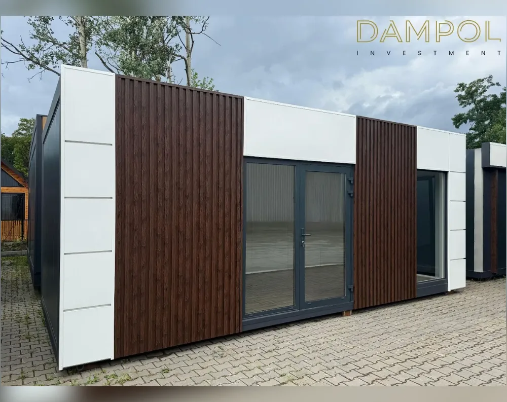 This modern modular unit has a long, low rectangular shape with a flat roof, viewed from an angled corner. The exterior alternates between smooth white paneling and vertical dark wood-toned slat cladding, creating a striped, contemporary look. The white sections feature subtle horizontal seam lines for a sleek finish. Centered on the front face is a dark-framed double glass door with a silver handle, flanked by wood-slat panels. To its right sits a large fixed rectangular window, also dark-framed, matching the door's frame color. The structure sits on paved stone ground, with trees and a cloudy sky visible behind it, and another similar unit partially visible to the right.