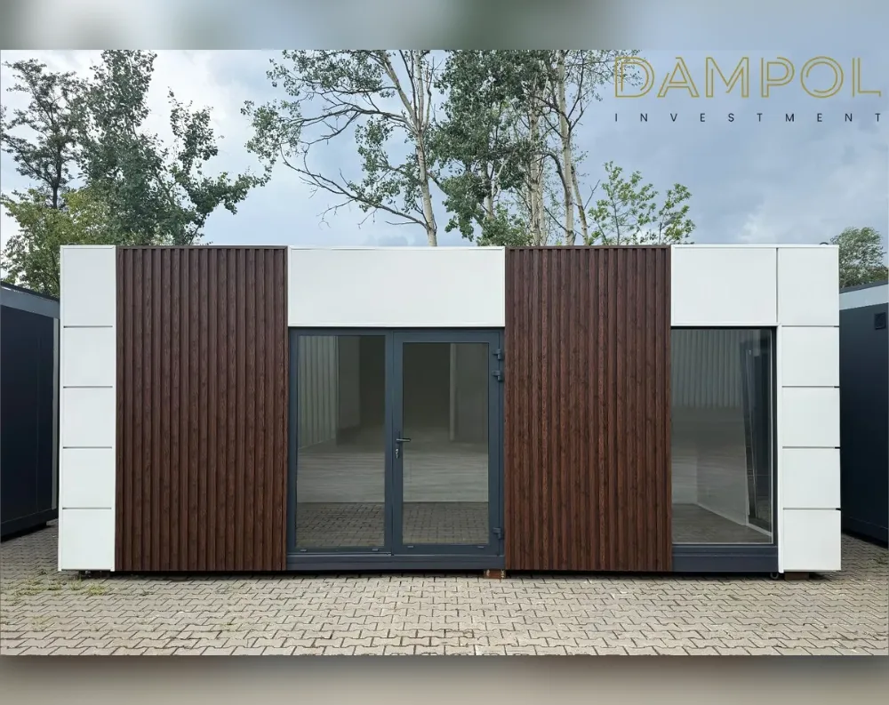 This modern modular building has a wide, low-profile rectangular shape with a flat roof. Its façade alternates between panels of dark vertical wood-slat cladding and smooth white paneling, creating a striking contrasting rhythm across the front. At the center sits a dark-framed glass double door, flanked on either side by large fixed windows set into the wood-paneled sections. The upper band above the entrance is finished in clean white, emphasizing the horizontal lines of the structure. The building rests on a paved brick surface, with mature green trees and a cloudy sky forming the backdrop, giving it a sleek, contemporary garden-office aesthetic.