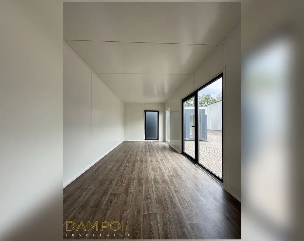 This photo shows the interior of a modular building, a long, narrow rectangular room with a clean, minimalist finish. The walls and ceiling are smooth white panels, divided by faint seam lines that hint at the modular construction. Dark wood-toned laminate flooring runs the length of the space, adding warmth against the white backdrop. At the far end, a black-framed glass door sits on the left, while to the right, a large black-framed sliding glass door and adjacent window fill the wall, letting in daylight and revealing a paved courtyard and another gray building outside. The proportions feel spacious yet efficient, with no visible furniture, emphasizing the room's clean, functional design.