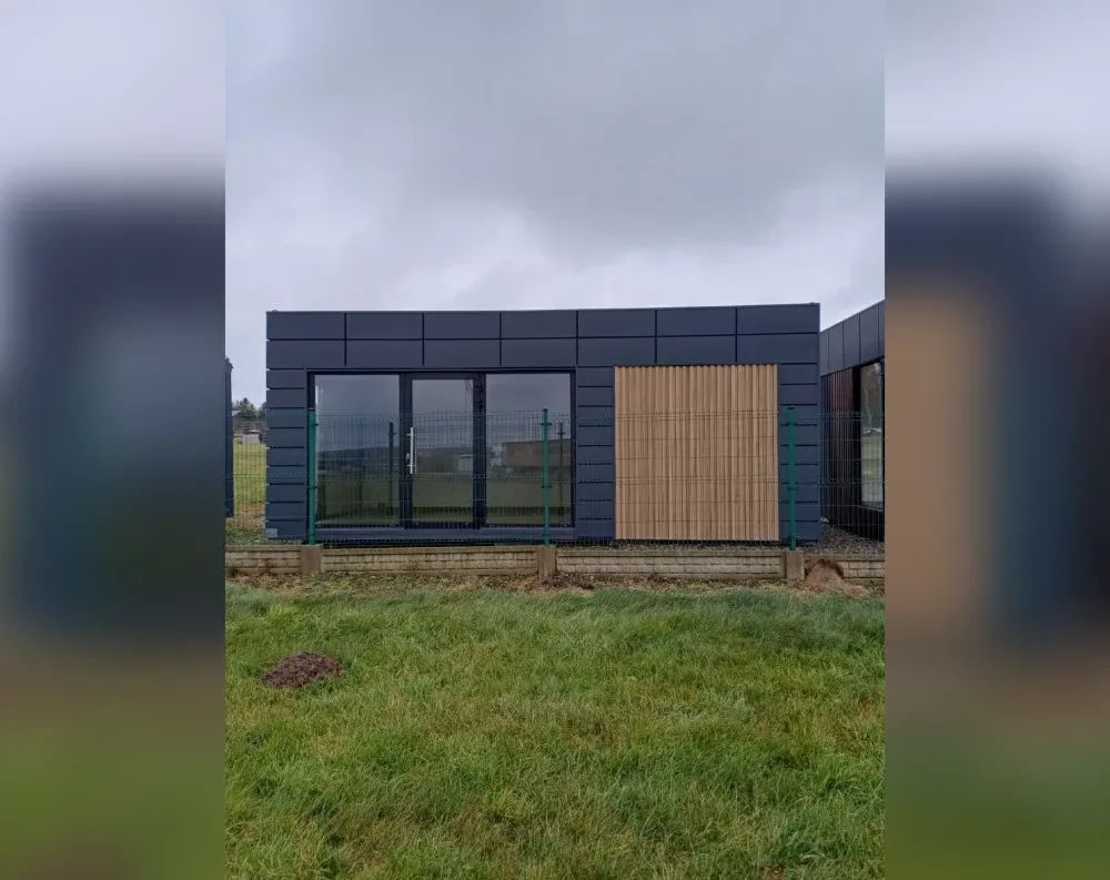This modern modular building sits low and horizontal, with a flat roofline and dark charcoal-grey metal cladding arranged in rectangular panels across its facade. The front features a large glazed double door with slim black frames positioned left of centre, offering full-height glass panels that reveal glimpses of the interior and landscape beyond. To the right of the entrance, a striking vertical wood-slat panel in warm honey-toned timber creates a natural accent against the dark exterior. Slender green metal fence posts stand in front of the building, hinting at an unfinished perimeter fence. The structure rests on a light concrete foundation strip, surrounded by patchy raw earth and unmown green grass in the foreground, under an overcast grey sky.
