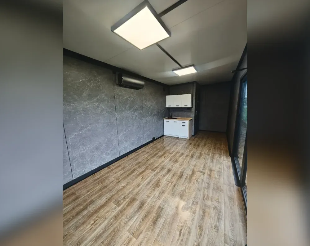 This is an interior view of a compact modular office or garden room, showing a single open room with a clean, modern finish. The floor is laid with warm honey-toned wood-effect laminate planks running the length of the space. One wall is finished in a dark grey marbled panel cladding, contrasted by plain pale ceiling panels fitted with two flush LED light panels. A small kitchenette sits along the back wall, featuring white upper and lower cabinets, a stainless sink, and a wall-mounted black unit above (likely an air conditioning system). To the right, a dark-framed glass sliding door and adjoining window let in natural light, with a solid door further along the same wall.