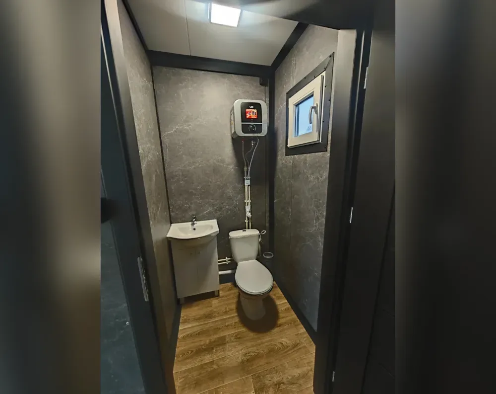 This compact modular unit houses a small bathroom, viewed through a dark-framed doorway that swings open to reveal the space beyond. The interior walls are clad in dark grey marbled paneling, giving a sleek, polished appearance, while the floor features warm honey-toned wood-effect flooring. A single small square window with a dark frame sits on the right wall, fitted with a fold-out handle. Inside, a white toilet and compact wall-mounted sink with a cabinet base occupy the space, alongside an electric water heater mounted high on the back wall with visible pipework running down. The ceiling holds a recessed square light fixture, and the trim throughout is finished in matching dark brown-black tones.