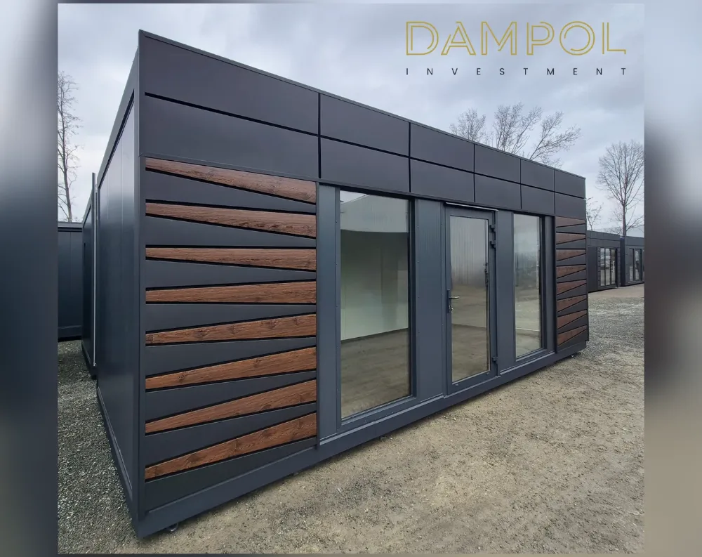 This modular building has a clean, rectangular footprint with a low-pitched, slanted roof that slopes gently from front to back. The exterior cladding is finished in a matte charcoal-black panel system, accented by two vertical sections of warm brown wood-look horizontal slats positioned on the front corners, framing the entrance. The front façade features a central glass door flanked by two large fixed windows, all set in dark anthracite frames, allowing full visibility into the empty interior space. The structure sits on a gravel surface, with similar modular units visible in the background, suggesting an outdoor showroom or storage yard setting.