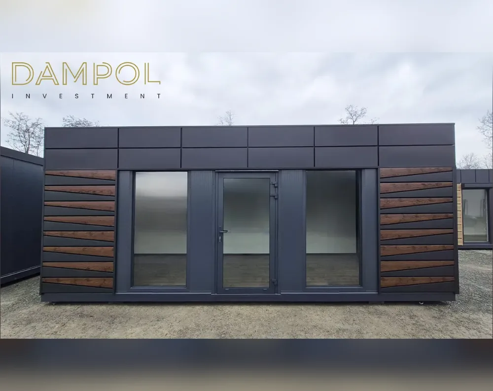 This modular building is a long, low, rectangular structure with a flat roof, giving it a sleek, contemporary profile. The exterior cladding is finished in a dark charcoal-grey panel system, smooth and matte across the upper band and lower base. Flanking each side of the entrance are vertical accent panels of warm brown wood-effect slats arranged in a chevron, diagonal pattern, adding warmth and texture. Centered on the façade is a dark-framed glass door flanked by two large fixed glass window panels, one on either side, allowing full-height visibility into the interior. The overall proportions are wide and horizontal, with clean lines, a minimalist aesthetic, and a raised base sitting slightly above the gravel ground.