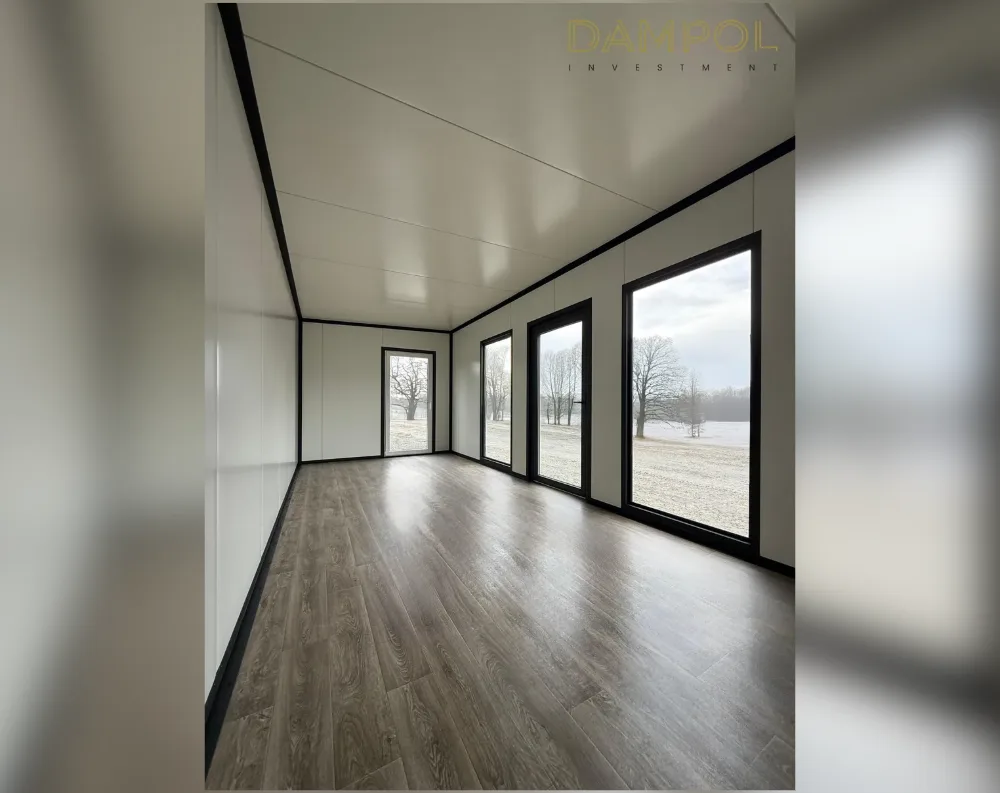 This is the interior of an empty modular building, viewed down its length toward a far wall lined with glazing. The space is narrow and elongated, with a flat white ceiling and matching pale wall panels framed by dark, almost black trim strips that outline every joint and edge. Along the right-hand wall stand three tall floor-to-ceiling windows with black frames, spaced evenly apart, looking out onto a snowy field dotted with bare trees. At the far end, a narrower black-framed glass door sits centered in the end wall, offering another view outside. The flooring is a glossy dark wood-look laminate, running diagonally and reflecting the light from the windows.