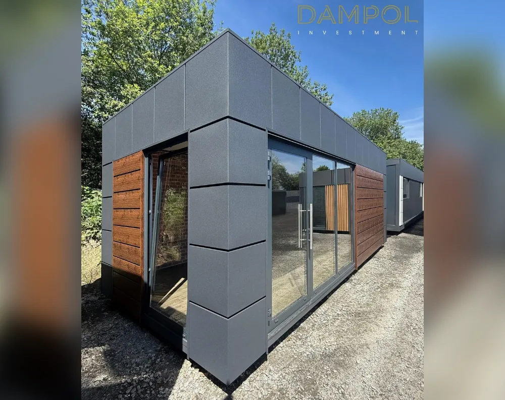 This modular garden building has a sleek, rectangular shape with a flat roof and clean, minimalist lines. The exterior is clad in dark charcoal-grey panels with subtle vertical seams, contrasted by warm horizontal wood-toned slats framing the entrances. At the front corner, a fully glazed double door system sits recessed within the wood paneling, opened to reveal the interior. Adjacent to it, a large sliding glass door with slim black frames spans much of the front façade, offering an expansive view inside. The structure sits on a gravel surface, with greenery and trees surrounding it, and a matching secondary unit is visible in the background.