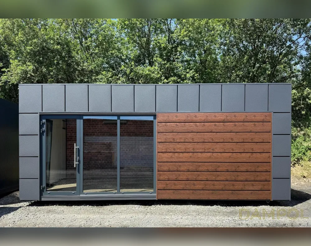 This modular garden building is a long, low rectangular structure with a flat roof, sitting on a gravel base against a backdrop of dense green trees. The left two-thirds of the facade are clad in textured grey composite panels arranged in a grid pattern, while the right third features horizontal wood-effect paneling in a warm brown tone. A large glass system dominates the left side, combining a hinged door with sliding glass panels, all framed in dark grey metal, offering a clear view into the brick-walled interior. The structure's clean lines, contrasting materials, and minimalist design give it a modern, functional aesthetic suited for use as an office or studio space.