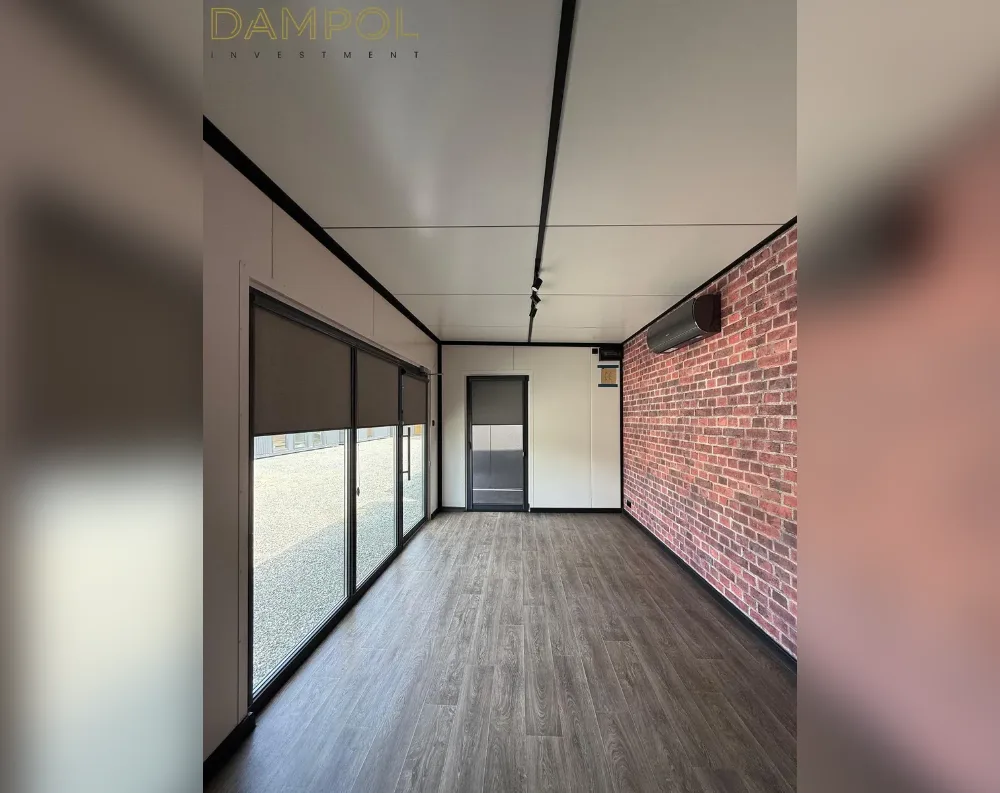 This is an interior view of a modular garden office with a rectangular, elongated layout. One long wall features a full-length glass sliding door system with dark frames and retractable brown roller blinds, offering natural light and outdoor views onto a gravel area. The opposite wall showcases exposed red brick accenting, paired with a dark wall-mounted air conditioning unit. The ceiling is flat, white paneled, with visible black seam lines running its length. A single dark-framed interior door sits centrally along the back wall. Flooring is medium-toned wood-effect laminate, and the overall palette combines neutral white walls with brick and black trim accents for contrast.