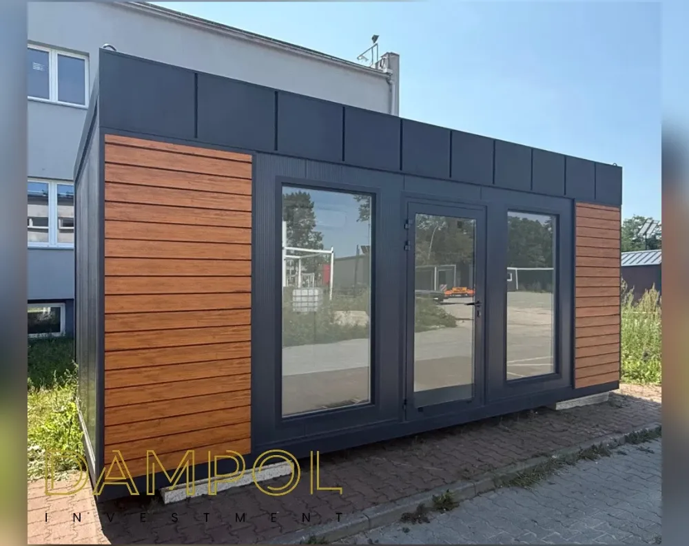 This modular garden office is a sleek rectangular structure with a flat roof, elongated in shape to suggest a spacious single-room interior. The front façade combines two contrasting cladding materials: warm honey-toned horizontal wood-effect paneling on both outer sections, framing a central dark charcoal-grey area finished in smooth vertical panels. This dark central section houses the entryway—a full-height glass door flanked symmetrically by two large fixed glass windows, all set in matching anthracite frames that let in abundant light and reflect the surrounding trees and pavement. The roofline and upper wall are finished in the same dark grey paneling, creating a clean, modern contrast against the wood accents below.