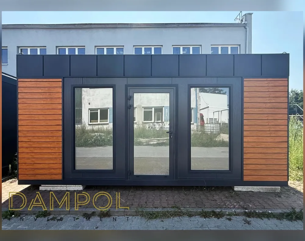 This modular garden office is a low, rectangular structure with a flat roof, wider than it is tall, giving it a sleek horizontal profile. The facade is dominated by a dark charcoal-grey panel cladding, contrasted by two vertical sections of warm honey-toned wood-effect slats bookending each side. Centered on the front are two large fixed glass windows flanking a full-height glass entry door with a slim black frame and handle, all reflecting the surroundings. Above, a paneled dark fascia band runs the width of the roofline. The structure rests on simple concrete block footings, set on a paved brick surface.