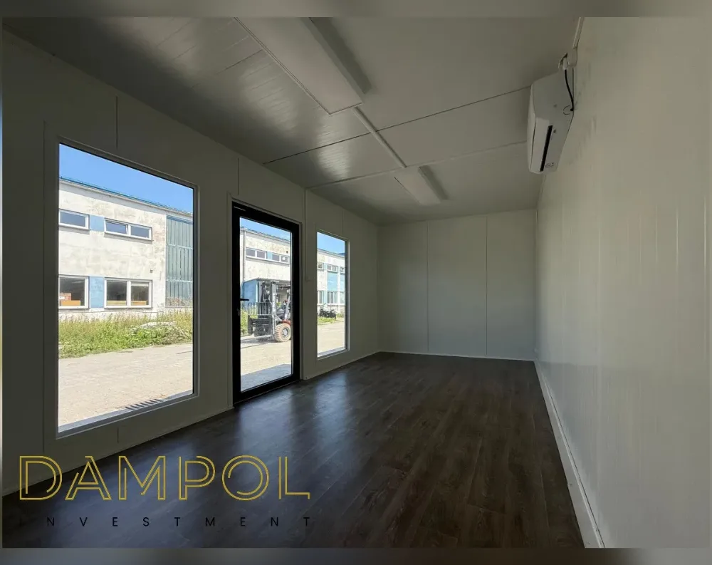 This is an interior view of a modular office unit with dark laminate flooring and pale, panelled walls and ceiling. On the left side, two large fixed windows flank a central glass-panelled door with a black frame, all offering a view of an outdoor industrial yard with another building and a forklift visible outside. The ceiling is flat with recessed linear lighting strips running across it, and a wall-mounted air conditioning unit sits in the upper right corner. The walls are finished in smooth, light beige or off-white panels with visible seams, giving a clean, minimalist look. The room appears rectangular and elongated, with simple, functional proportions and no additional furnishings visible.
