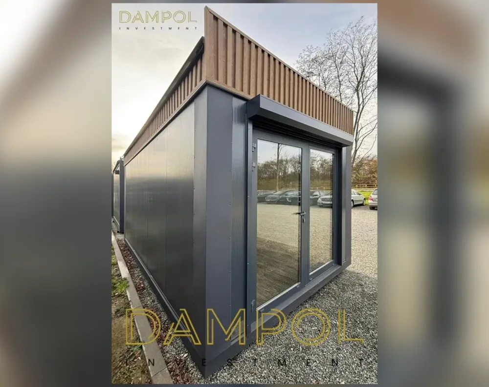 This modular garden office is a sleek, rectangular structure with a low-pitched flat roof topped by vertical wood-toned slatted panels running along the upper edge and wrapping down one corner. The main body is clad in dark charcoal-grey ribbed metal panelling, giving it a modern, streamlined appearance. The front façade features a full-height glass double door with slim dark frames, positioned right of center, paired with a matching glass side panel to its left, allowing ample natural light inside. The structure has clean, sharp edges and a boxy yet elegant silhouette, sitting on a gravel surface with trees and parked cars visible in the background.