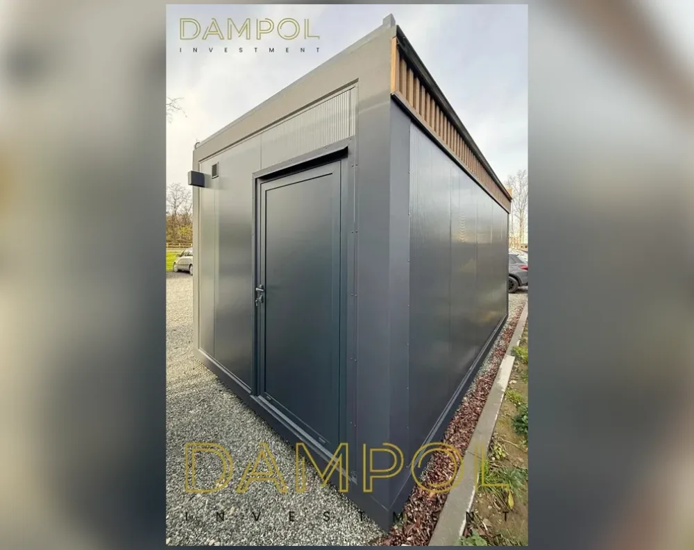 This modular garden building has a sleek, rectangular shape with clean, boxy proportions, appearing single-story with a slightly elevated flat roof edge. The exterior cladding is finished in dark charcoal-grey panels with a smooth, ribbed texture, giving it a modern, industrial look. A single solid door sits centrally on the front-facing wall, painted the same dark grey to blend seamlessly with the walls. To its left is one small rectangular window, positioned high on the wall for privacy and light. Along the upper edge of the structure runs a band of natural wood-toned slats, creating a warm contrasting accent beneath the flat, slightly overhanging roofline. The overall impression is minimalist, contemporary, and finished with precision.