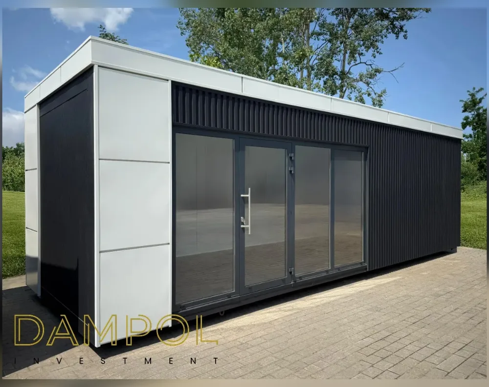 This modern modular structure has a long, low rectangular form with a flat, overhanging roof edged in white trim. The main facade is dominated by a wide glass frontage divided into four panels—two central hinged doors with a slim metal handle, flanked by fixed glass sections—all set in dark charcoal frames. Above this glazing, ridged black vertical cladding wraps across the upper wall and continues along the building's side, contrasting with smooth white panels on the near end wall. The roofline is crisp and minimal, giving a sleek, contemporary silhouette. The unit rests on a paved surface, with green lawn and trees surrounding it, framed by a bright blue sky.