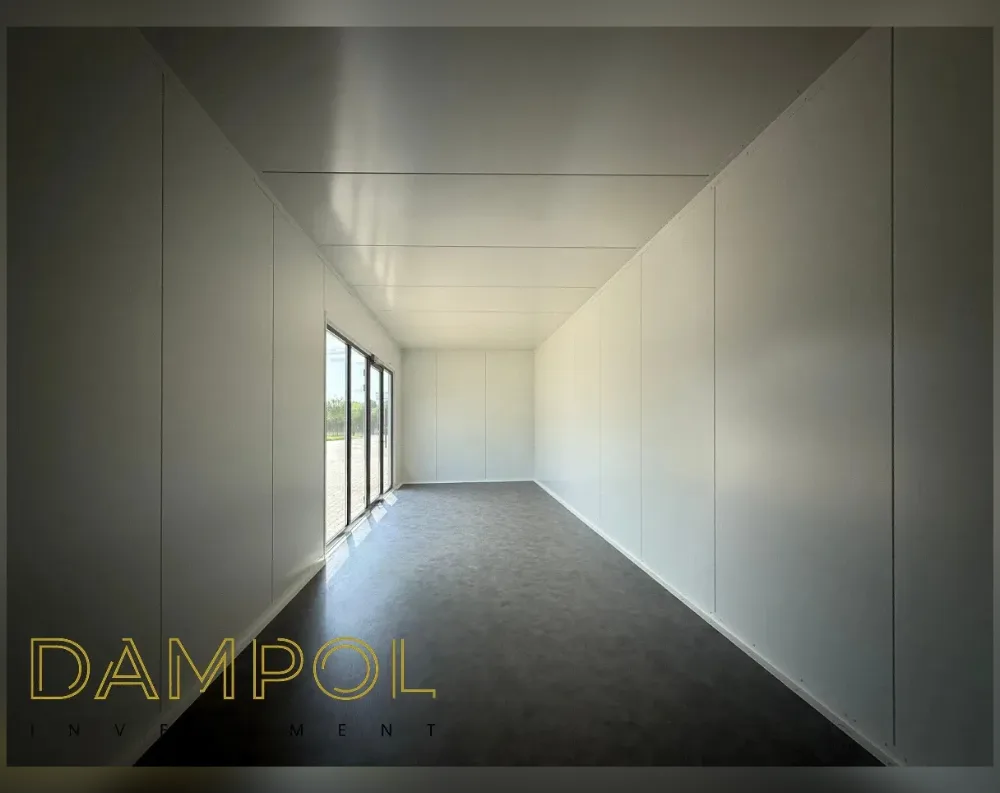 This photo shows the interior of a long, narrow modular room with a simple rectangular footprint and a flat ceiling. The walls and ceiling are finished in smooth white panels, divided by subtle seam lines that run horizontally and vertically. At the far end of the room, on the left-hand wall, sits a tall sliding glass door with slim dark frames, divided into multiple panes, allowing daylight to spill across the dark grey flooring. The floor has a speckled, matte finish. No additional windows or doors are visible beyond this single glazed entry point. The space feels minimalist, bright, and clean, with strong linear perspective drawing the eye toward the glass door.
