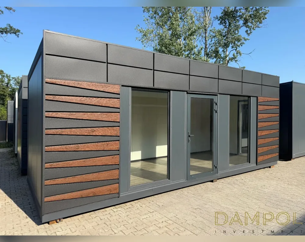 This modular garden office is a sleek, rectangular single-story structure with a flat roof and clean, elongated proportions. The dark charcoal-grey cladding forms the base, accented by two diagonal wood-toned panel sections flanking the entrance, their angled slats adding warmth and texture. The front façade features a central glass double door flanked by two large fixed glass panels, creating a bright, symmetrical entrance with three see-through sections total. Above, a paneled grey fascia band wraps the upper portion beneath the flat roofline. The structure sits on a paved stone surface, with greenery and trees in the background beneath a clear blue sky, giving the building a modern, minimalist aesthetic.