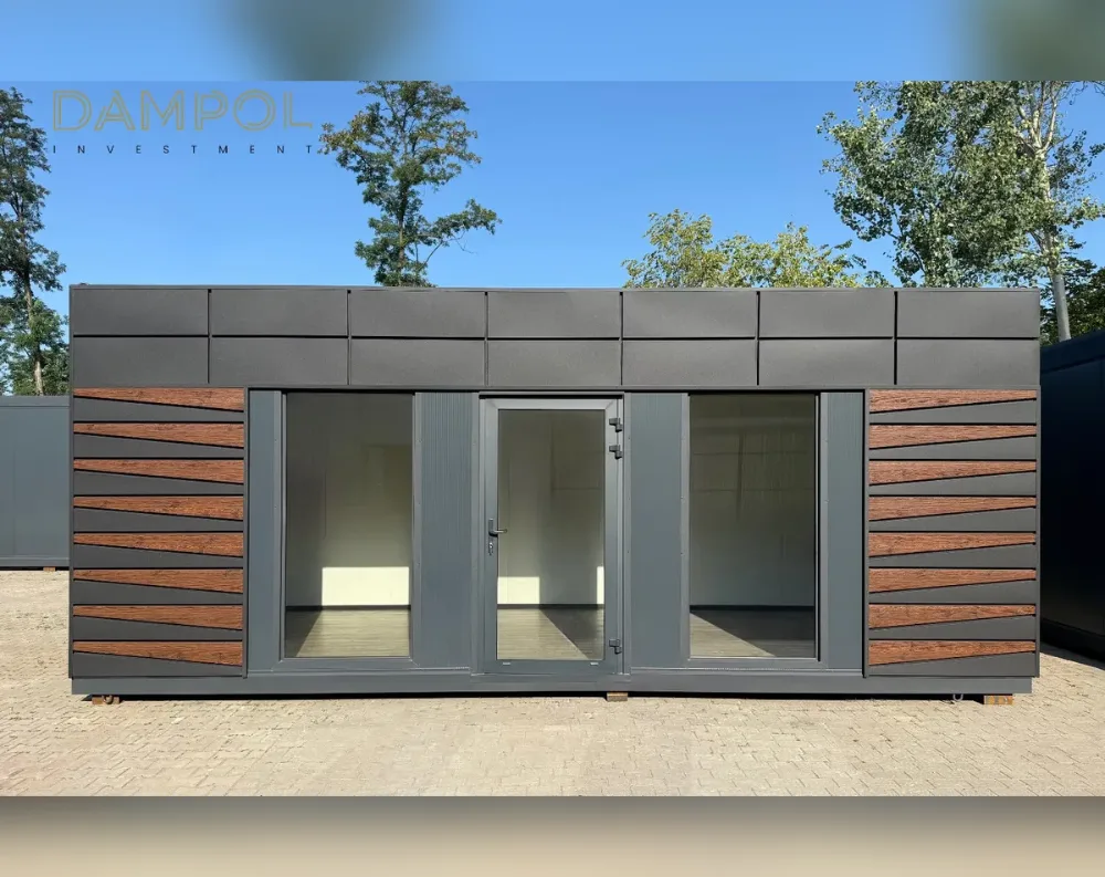 This modular garden office presents a long, low rectangular silhouette with a flat roof, its dark charcoal-grey paneled fascia forming a bold horizontal band across the top. The main facade combines matte dark-grey cladding with striking diagonal wood-toned slat accents on both outer sections, creating a herringbone-like pattern. Centered on the front are two large fixed glass window panels flanking a central glazed double door with slim vertical panes, all framed in matching dark-grey trim. The structure sits on small support blocks above a paved surface, with trees and blue sky visible behind it, giving the building a sleek, modern, cabin-like appearance with clean lines and contrasting textures.