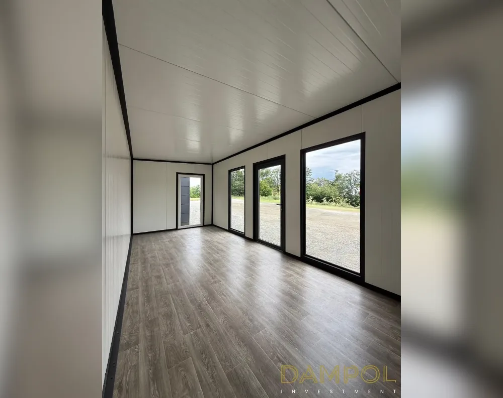 This is the interior of a long, narrow modular room with a flat white ceiling and matching pale walls, all trimmed in black framing that outlines every edge and joint. The vinyl flooring has a warm grey-brown wood-grain finish, running the length of the space. Along the right-hand wall are three tall black-framed windows, evenly spaced, letting in daylight and views of gravel ground and greenery outside. At the far end, a black-framed glass door sits in the corner, offering an additional exit. The layout is simple and rectangular, with clean structural lines, a low-pitched ceiling, and an open, uncluttered feel throughout the entire room.