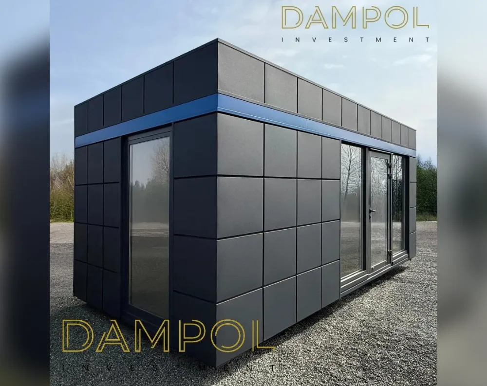 This modern modular building sits on a gravel surface, presenting a sleek rectangular box shape with a flat roof. The exterior is clad in dark charcoal-grey square panels arranged in a grid pattern, giving it a crisp, contemporary look. A horizontal navy-blue accent stripe wraps around the structure just above the entrance, adding a pop of color. On the front-facing corner, there's a tall glass door with a slim frame, and beside it a large fixed window pane. Around the corner on the shorter visible side, another full-height glass panel serves as a window. The overall proportions are low and elongated, giving it a minimalist, cabin-like garden office appearance.