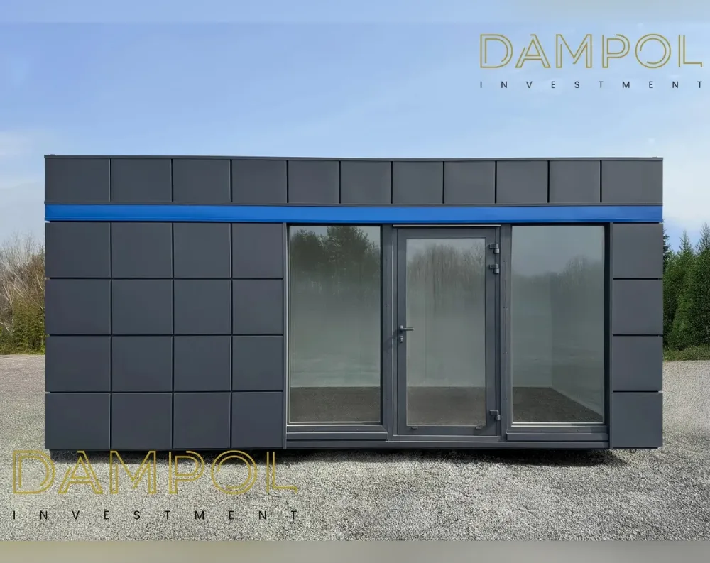 This is a sleek, rectangular modular unit with a flat roof, wider than it is tall, sitting on gravel ground with trees visible behind it. The exterior is clad in dark charcoal-grey paneling arranged in a grid pattern of square segments across the upper portion and left section. A bold blue horizontal stripe runs along the top edge, separating the paneled section from the glazed front. The right-of-center facade features a large glass and aluminum-framed entrance: a central hinged door flanked by two wide fixed glass panels, all set in dark grey frames, allowing a view straight through to the interior and greenery beyond. The structure appears mounted on a low base, giving it a slightly elevated, portable appearance.