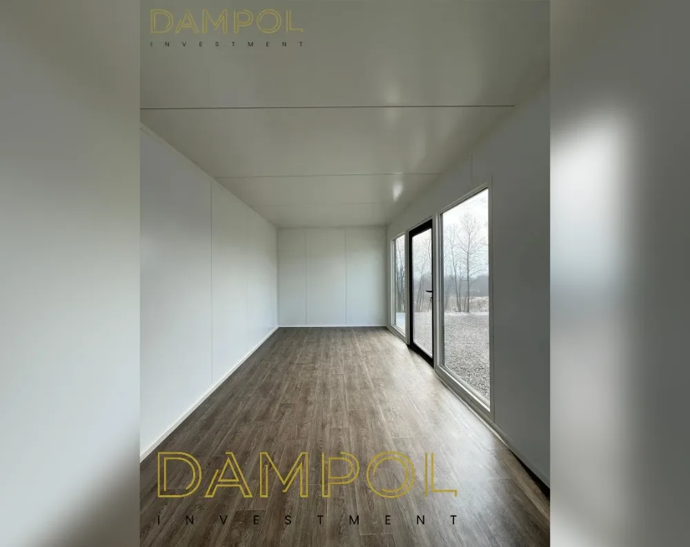 This modular room presents a long, narrow rectangular interior with clean, minimalist proportions. The walls are finished in smooth white paneling, seamlessly extending to a flat white ceiling with subtle linear seams marking the panel joints. At the far end, a full-height glass door with slender aluminum framing sits beside a matching tall window, both allowing abundant natural light to flood in and offering a view of bare trees and a gravel exterior. The flooring is a warm, medium-brown wood-look laminate with visible grain texture, running the length of the room and adding contrast to the white walls. The overall aesthetic is simple, bright, and open, with no visible roof detail from this interior angle.